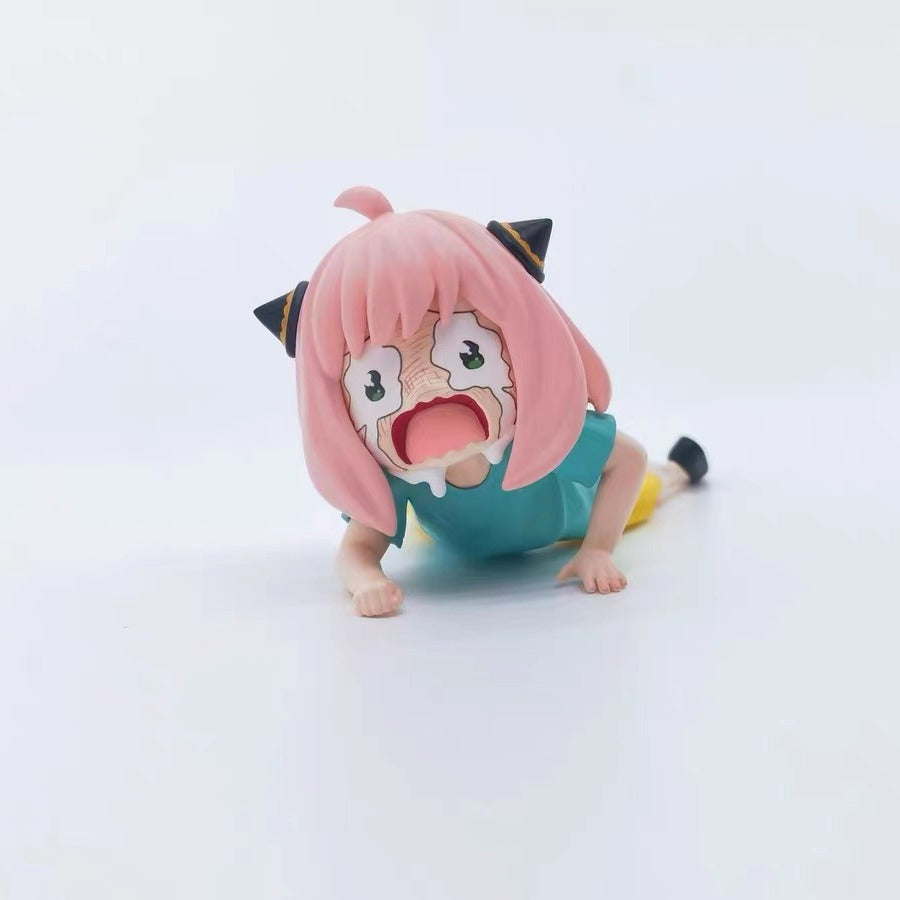 A 6cm PVC figure of Anya Forger from Spy × Family, showing her pink hair, wide eyes, and surprised open mouth, lies face down in a blue top and yellow shorts against a plain white backdrop.