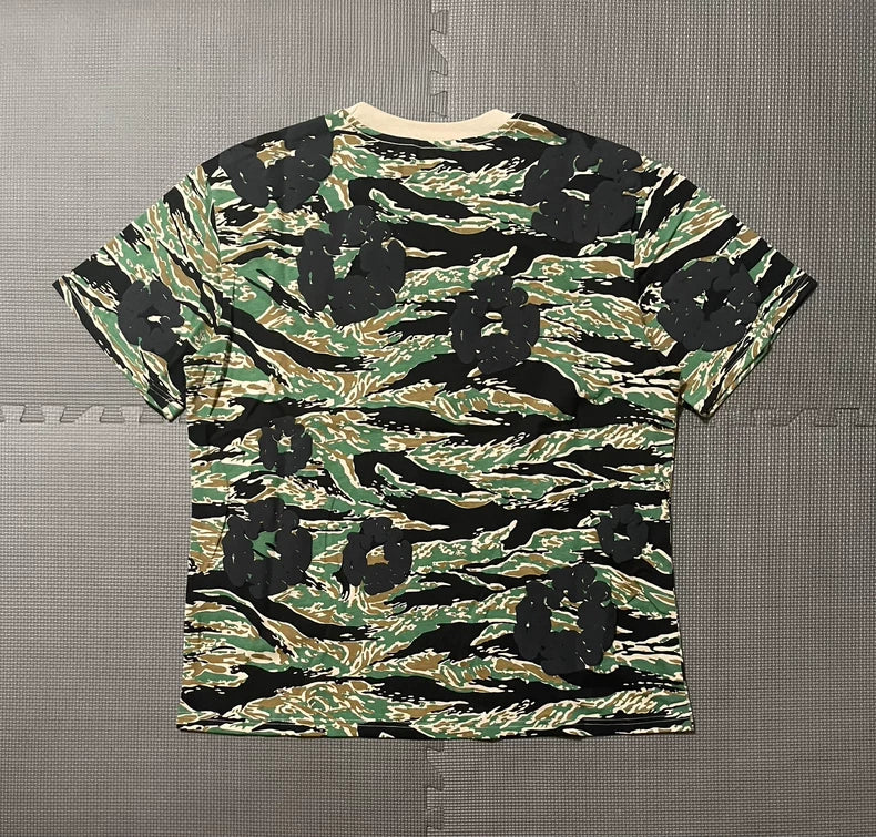 The Denim Tears Green Camo All-Over Print T-Shirt is a short-sleeved cotton tee featuring tiger stripe camouflage in green, brown, and black with bold black flower shapes, shown flat on a textured gray surface.