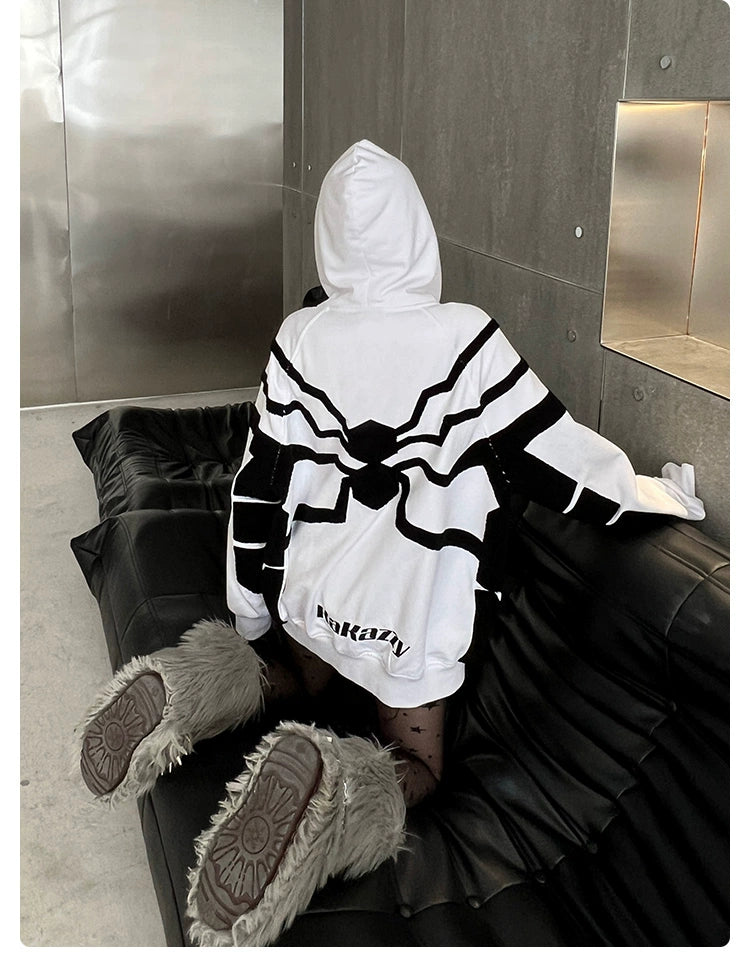 A person kneels on a black cushioned surface wearing a Seakoff Venom-Inspired White Hoodie, made from 400GSM cotton with a symmetrical black design on the back. Their furry gray boots and hood are up as they face a reflective metal wall.