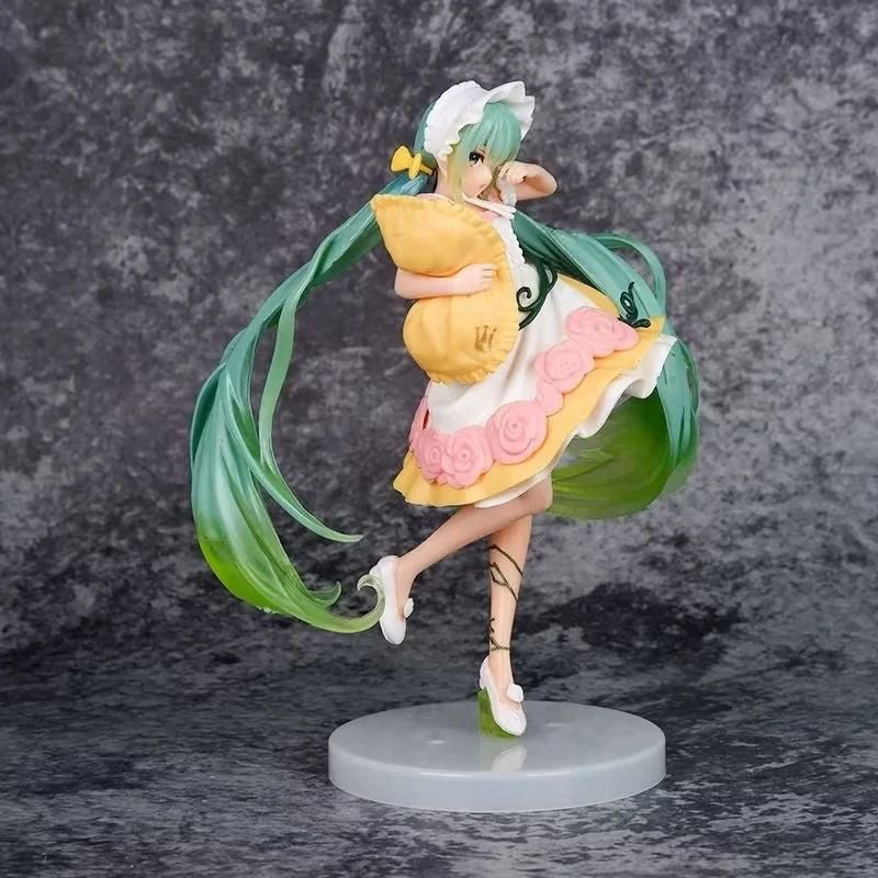 This Hatsune Miku Sleeping Beauty PVC Figure depicts the iconic Vocaloid with long green hair, a white bonnet and dress decorated with pink flowers, holding a yellow pillow. It strikes a dynamic pose on a gray round base against a textured backdrop. Collectible comes boxed and measures 20cm.