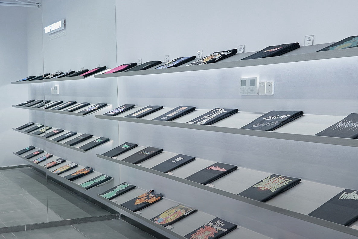A modern gallery with three long, slanted shelves displaying a series of open books. Each book has a unique cover design, and they are arranged neatly in rows along a white wall. The room has a minimalist aesthetic with gray flooring.