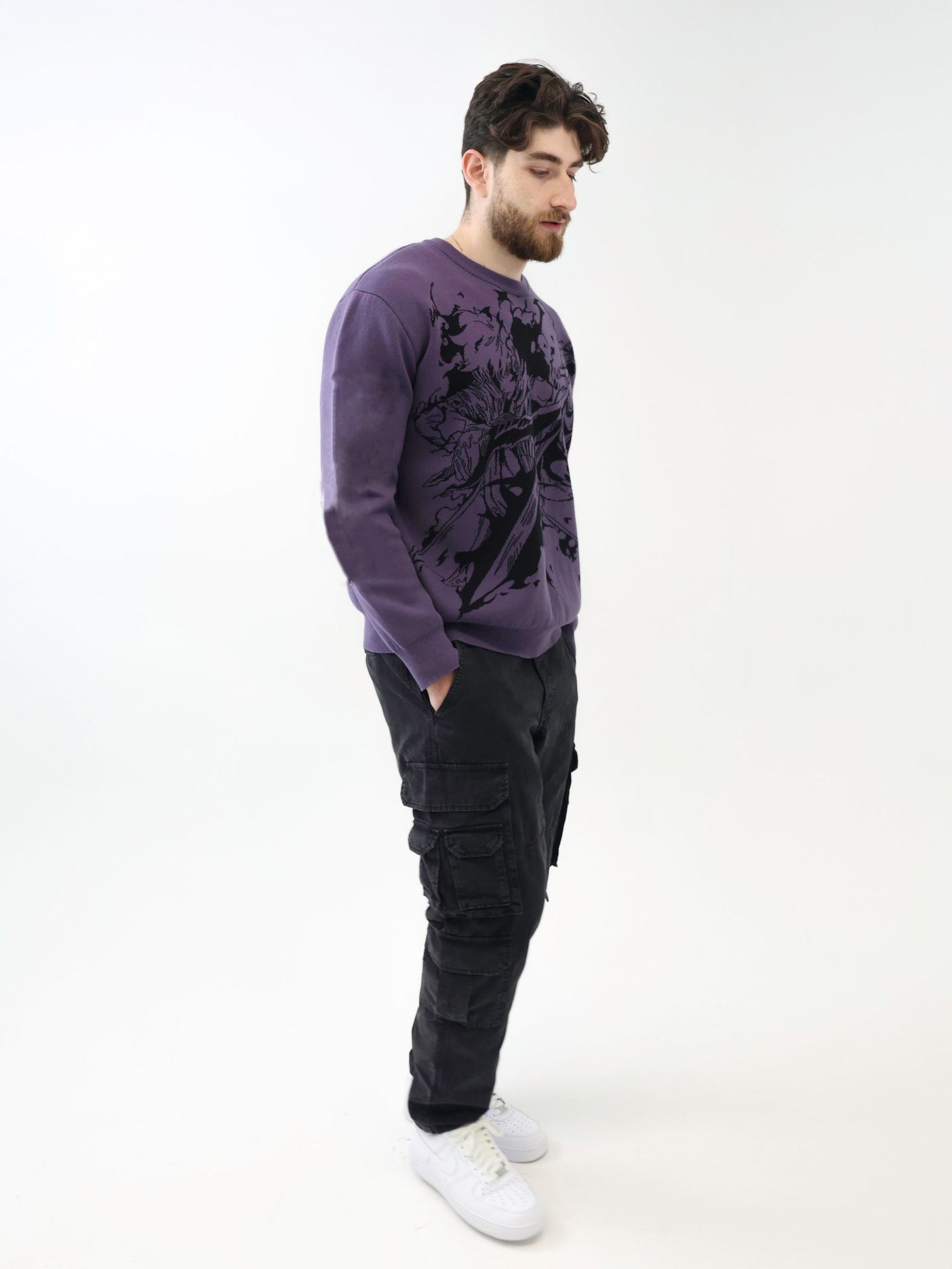 A bearded man in a purple Premium Solo Leveling-Inspired Knit Sweater with the Sung Jinwoo Shadow Monarch Jacquard Design pairs it with black cargo pants and white sneakers. He stands casually with hands in pockets against a plain white background.