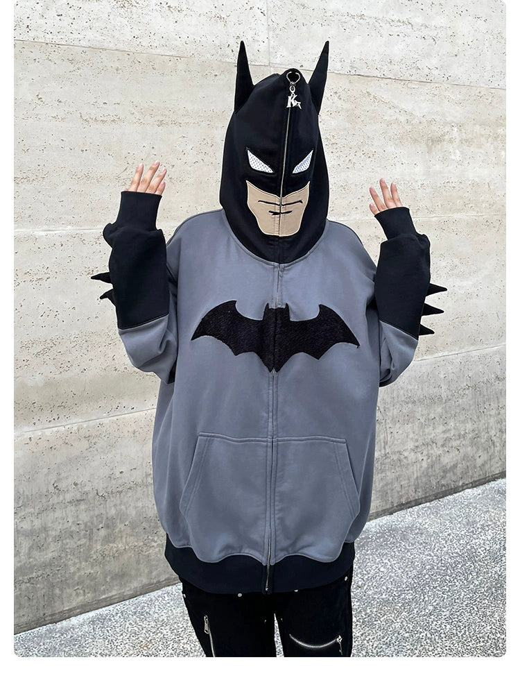 Someone wears a Seakoff Batman-Inspired Hoodie, an oversized grey and black zip-up sweatshirt with a superhero mask-style hood and black bat logo, featuring edgy spikes on the sleeves against a plain concrete wall.