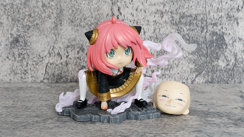 The Spy × Family Anya Forger PVC Figure features a chibi-style design with pink hair and black cat ears, crouching on a rocky base with swirling effects. It includes an interchangeable smiling head, all set against a textured gray background.