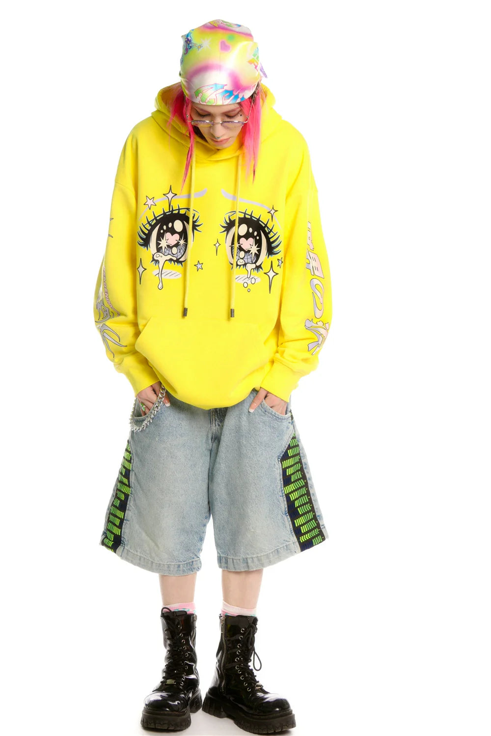A streetwear lover wears a Seakoff Kawaii Aesthetic Crying Eyes Hoodie featuring sleeve graphics. Paired with baggy denim shorts and black boots, their pink hair peeks from under a multicolored beanie. Glasses against the white background enhance the anime-inspired charm.