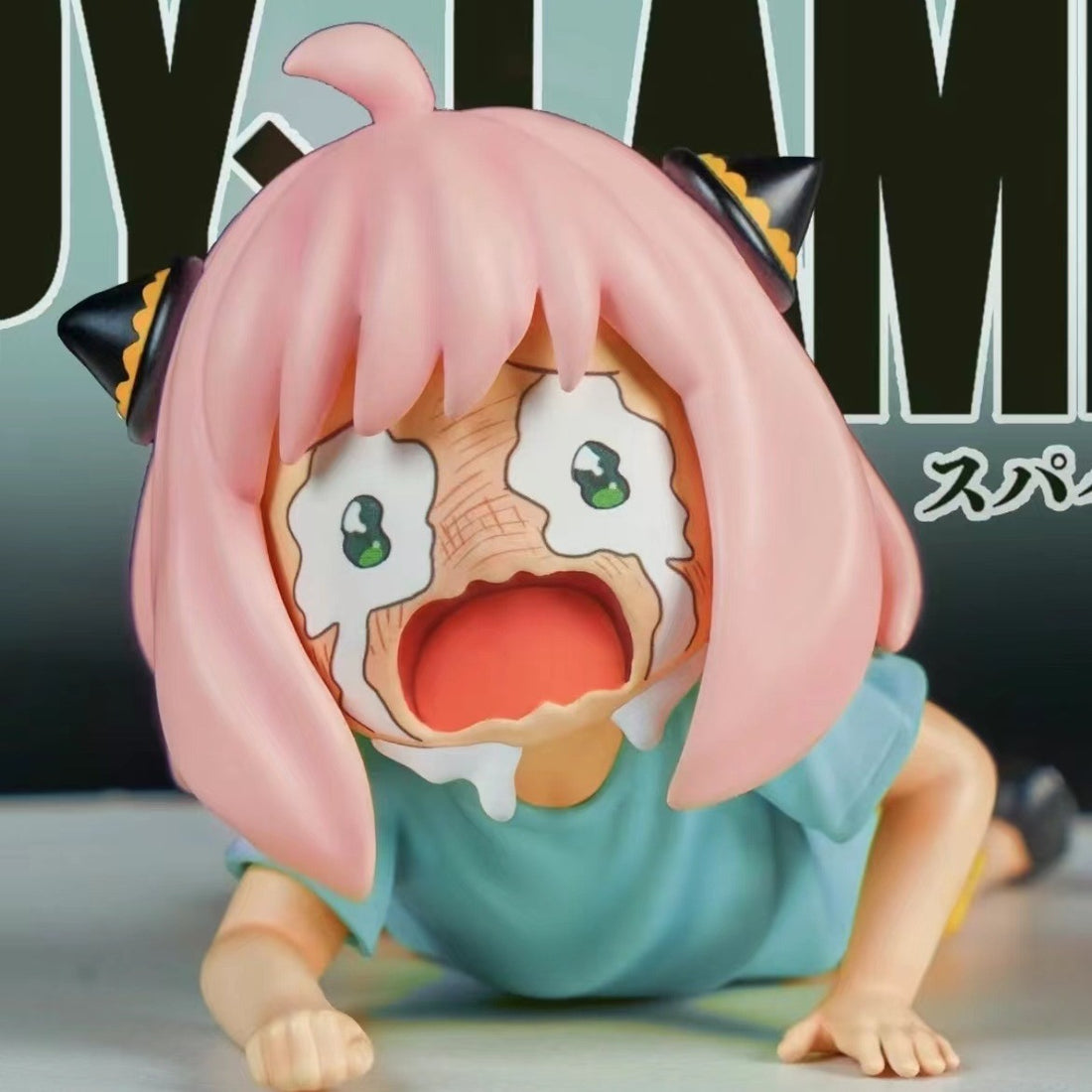 The Anya Forger Crying PVC Figure, a 6cm collectible from Spy × Family, features the chibi-style character on her hands and knees with pink hair and horn-like accessories. Her large eyes show an exaggerated crying expression amidst a background of partially obscured text.