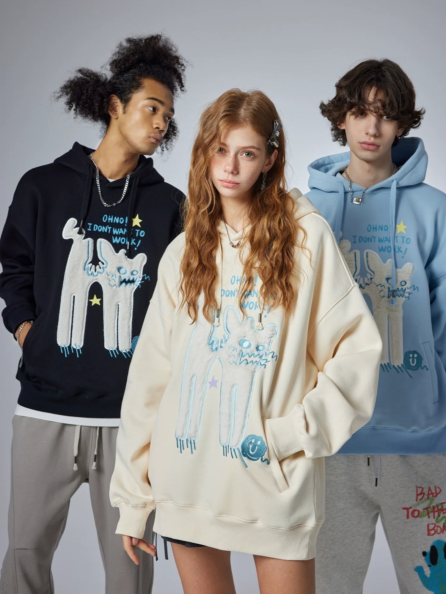 Three people model stylish oversized hoodies with cartoon designs. The left showcases a black hoodie, the middle features Seakoff's cream-colored "Oh No! I Don’t Want to Work!" hoodie with a cute lazy cat design, and the right wears blue. They're all posing against a neutral backdrop.