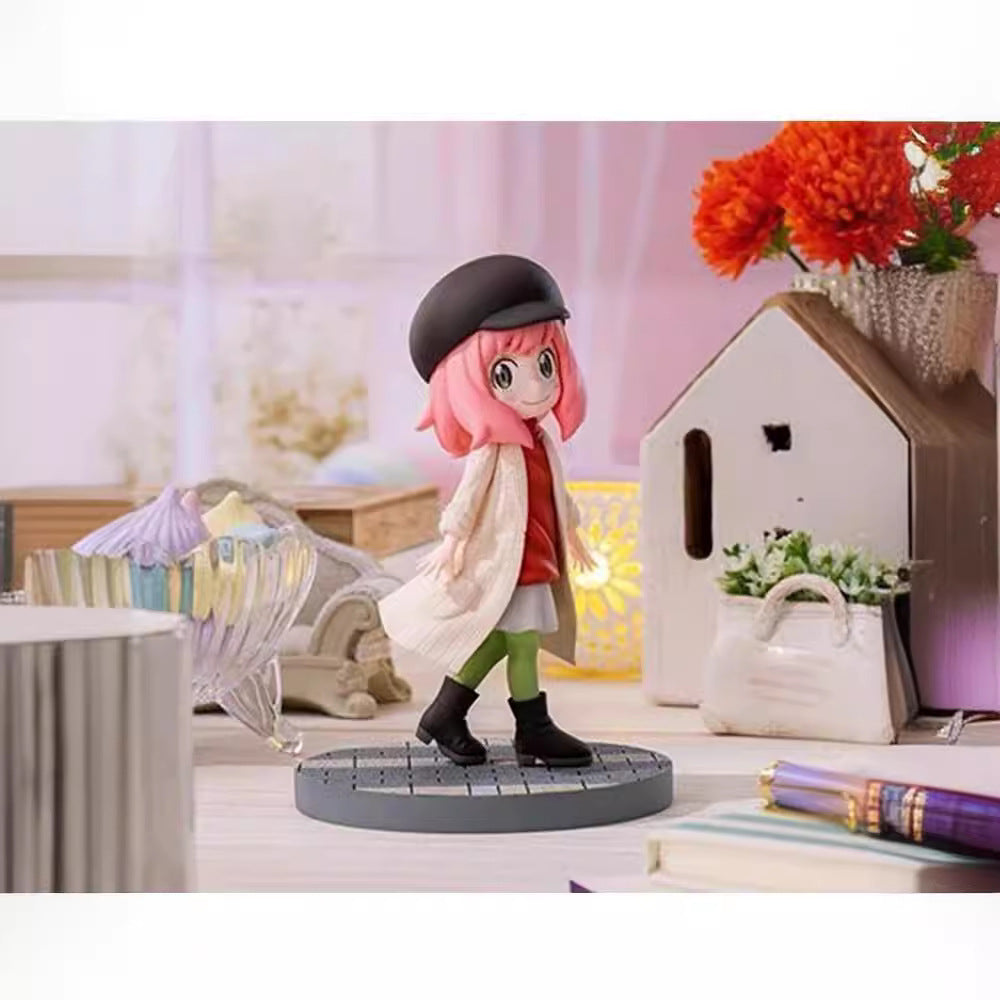 A 14 cm chibi-style Anya Forger figure from Spy × Family, wearing a pink hairstyle, black beret, white coat over a red dress with green leggings stands on a circular base. It includes an ornate glass bowl, books, house decor, and red flowers backdrop.