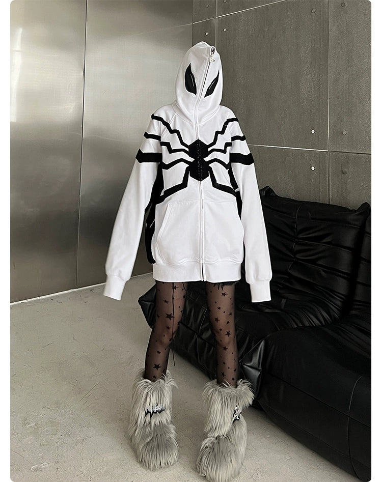 A person in a Seakoff Venom-Inspired White Hoodie with a black spider emblem, black star-patterned tights, and furry gray boots stands in an industrial-style room with metal and dark furniture. The hood of the 400GSM oversized superhero sweatshirt completely covers their face.