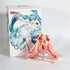 A PVC figure with long pink hair and cherry decorations sits before a box featuring anime-style art of Hatsune Miku with long blue hair. Both the figure and illustration share similar outfits and design, reflecting the playful style of the Hatsune Miku Cherry Sitting Pose 11cm Collectible.