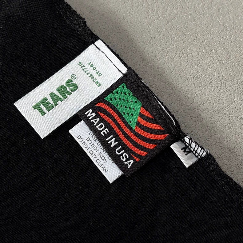 Close-up of tags on the Denim Tears Black Cotton Wreath All-Over Print T-Shirt. One tag shows Denim Tears®; another has a green and red American flag with MADE IN USA above care instructions on this oversized graphic tee.