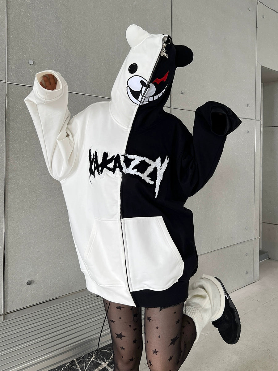 A person wears a large Seakoff Monokuma-Inspired Hoodie, a black and white zip-up featuring the iconic bear graphic, paired with star-patterned tights and black shoes. They pose against a light-colored wall—ideal for Danganronpa enthusiasts.