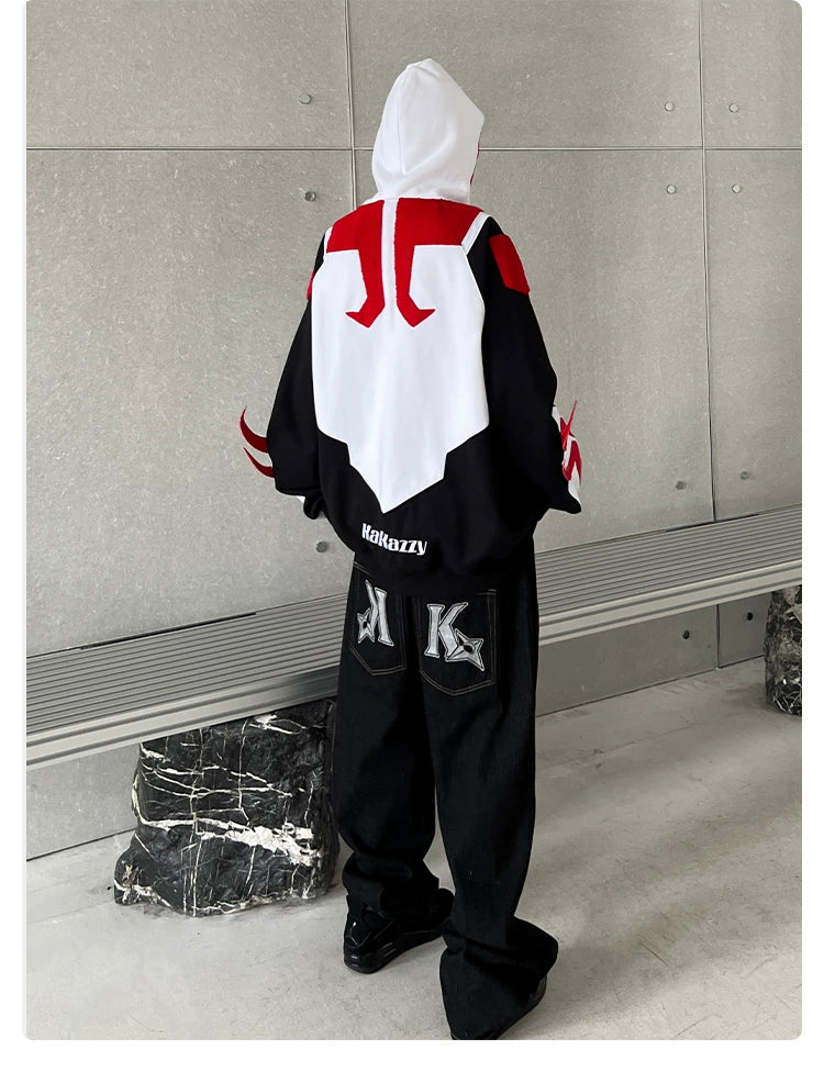 A person wearing a Seakoff Superhero-Inspired Hoodie—a 400GSM oversized black, red, and white zip-up streetwear sweatshirt with a large graphic on the back—faces a metallic wall. They also sport baggy black pants featuring a prominent letter &