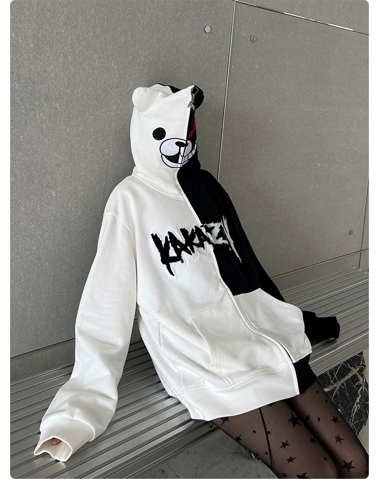 A person sits on a bench wearing a Seakoff Monokuma-Inspired Hoodie, an oversized black and white zip-up Danganronpa sweatshirt featuring a playful bear face and lettering. Paired with star-patterned tights, this anime hoodie stands out against the plain wall backdrop.