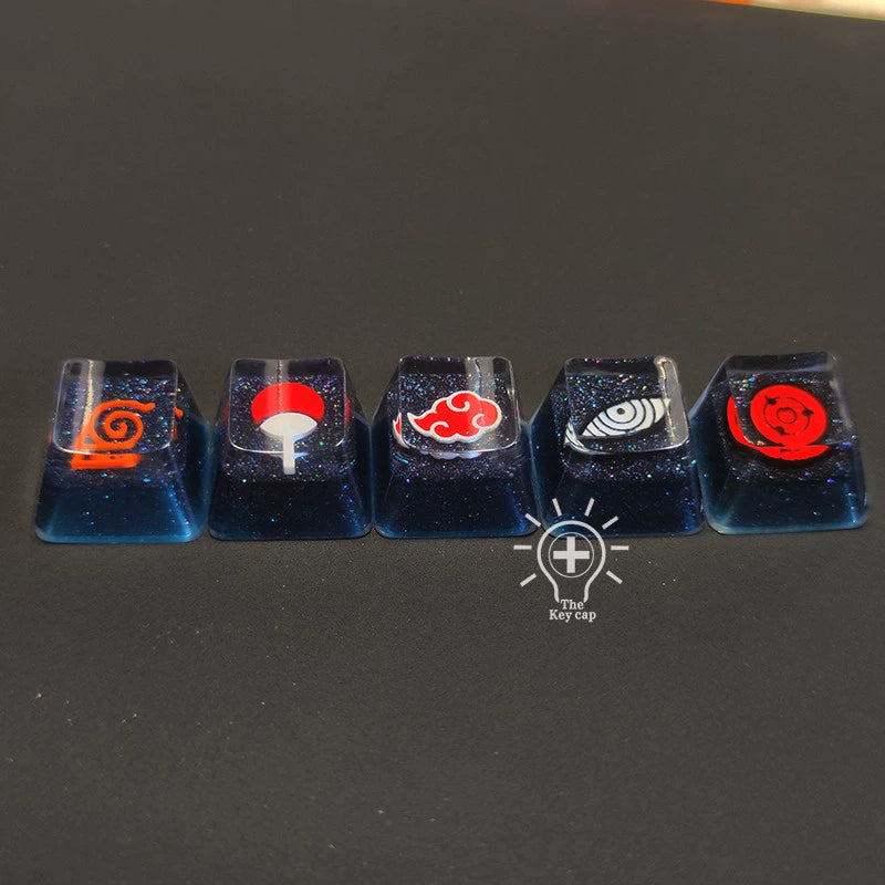 Five high-quality resin keycaps, each displaying different symbols from the Naruto Symbols Keycap Set—Hidden Village, Uchiha, Akatsuki—feature cosmic glitter designs with an illuminated effect against a dark surface.