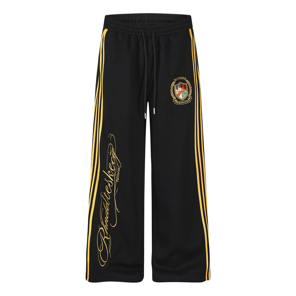 Black wide-leg track pants with three gold side stripes, embroidered crest badge at hip, and gold script graphic on front leg