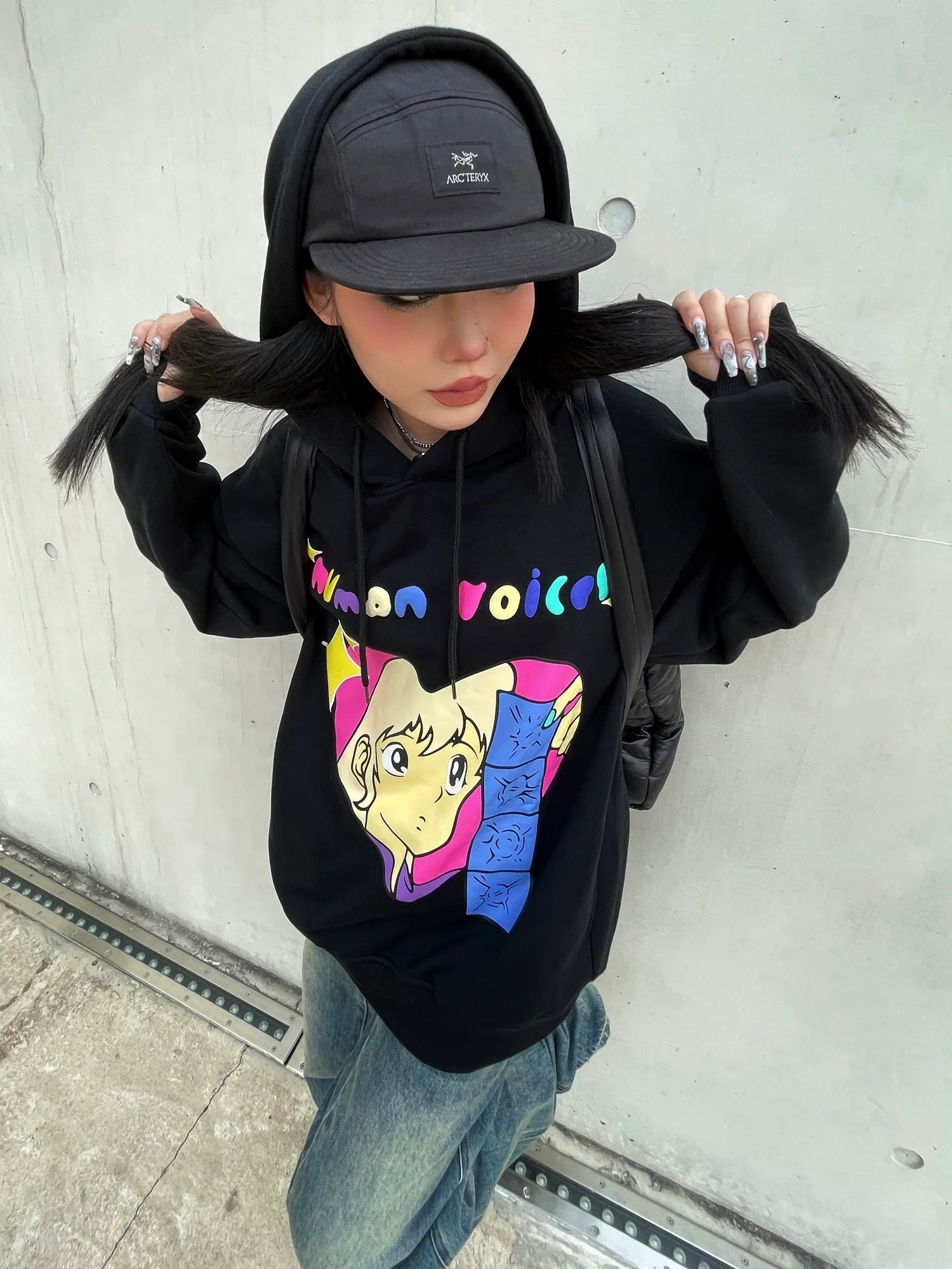 A person wearing a Seakoff Retro Anime Hoodie – Human Voices Graphic Pullover, jeans, and a black cap poses against a concrete wall. Holding pigtails with long painted nails, the hoodie's nostalgic graphic features pink and yellow elements.