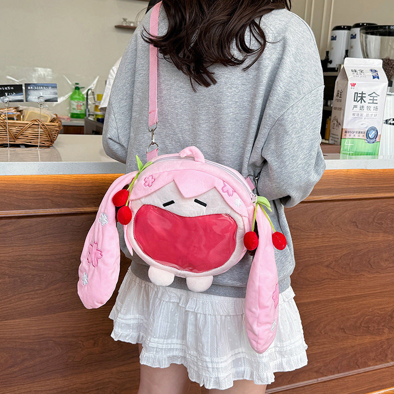 A person is standing in a room with tables and boxes, carrying a Seakoff Cute Anime Hatsune Miku Backpack. The plush ita bag features a pink cartoon character design, long ears, cherry decorations, and a transparent front pocket displaying anime merch. They are wearing a gray sweater and white skirt.