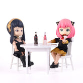 Two poseable Anya & Yor Forger Nendoroid figures from Spy × Family, one with black hair and gold accents (Yor) and the other with pink hair and cat ears (Anya), sit at a white table in black and gold outfits, with drinks between them.