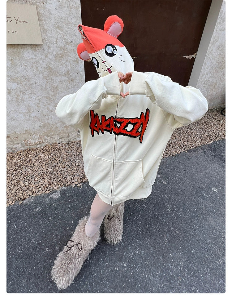 A person in a Seakoff white hoodie featuring red lettering and a cartoon mouse mask poses on the pavement. The outfit includes a red hat, beige pants, and fuzzy slippers, standing adorably in front of the white and brown wall—a perfect gift for any cartoon lover!.