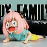 The Anya Forger Crying PVC Figure is a 6cm collectible from Spy × Family, featuring Anya with pink hair and large eyes in a crawling pose, wearing a blue shirt and yellow shorts. The background prominently displays "Spy Family.