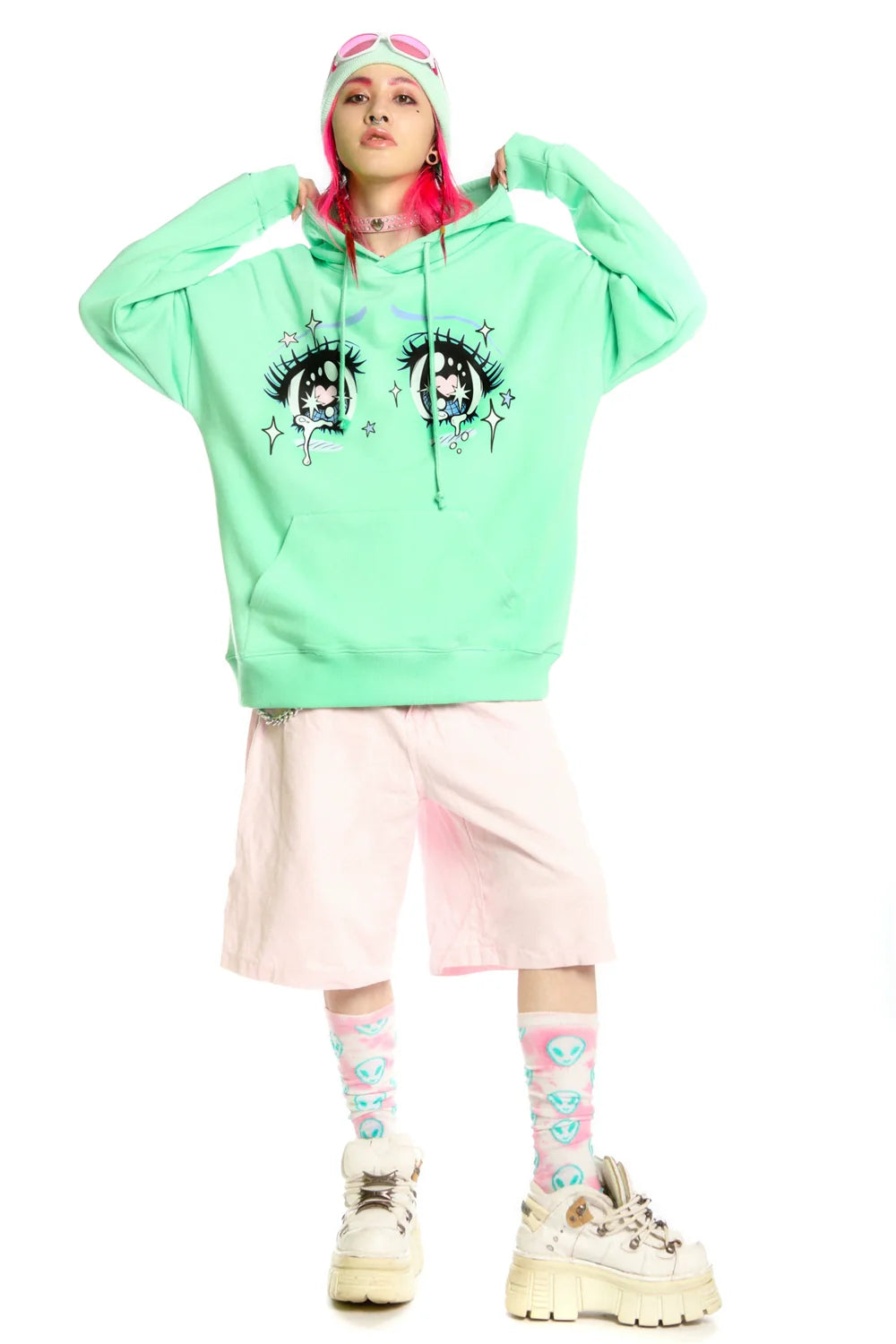 A person in Seakoff's mint green Kawaii Aesthetic Crying Eyes Hoodie with pastel anime graphics pairs it with light pink shorts and chunky sneakers. With pink hair, they're sporting pink goggles, a white and pink beanie, and blue alien socks against a white backdrop for streetwear vibes.