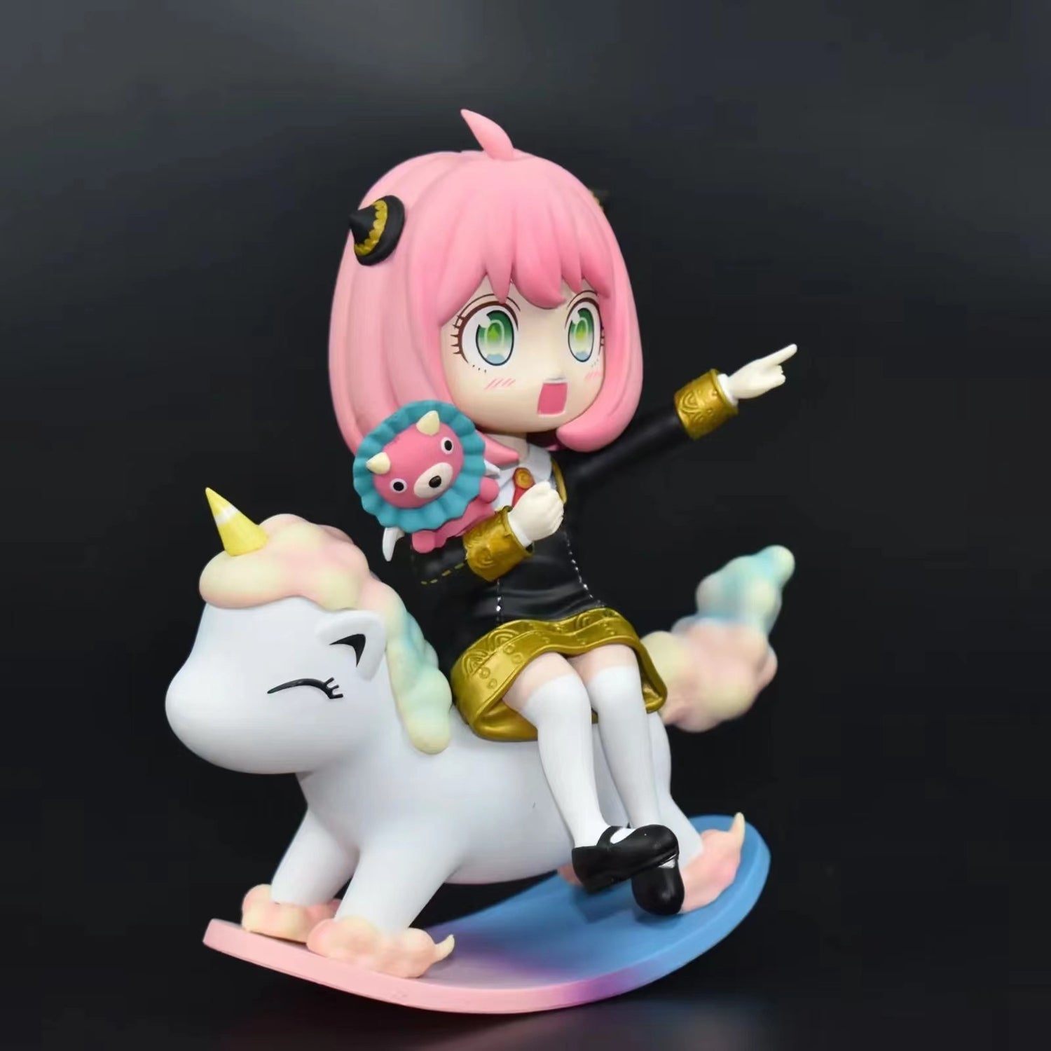 The Spy x Family Anya Forger PVC Figure, measuring 15 cm, showcases Anya with her pink hair and green eyes on a pastel unicorn rocking toy. She clutches a plush toy and playfully points to the side, set against a plain black background.