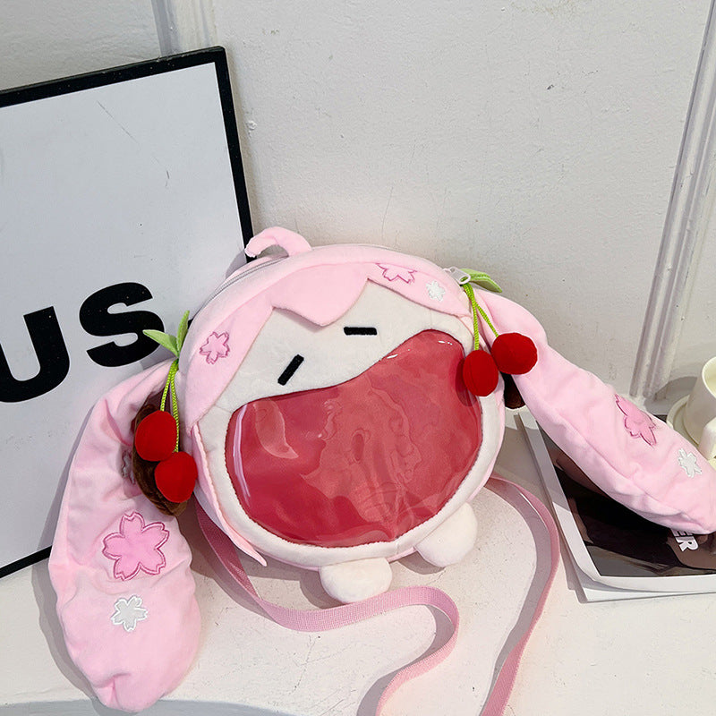 A Cute Anime Hatsune Miku Backpack by Seakoff, featuring a plush ita bag design with pink hair, cherry accessories, and a large open mouth. It has a detachable strap and is placed near a sign showing the letters &quot;US.