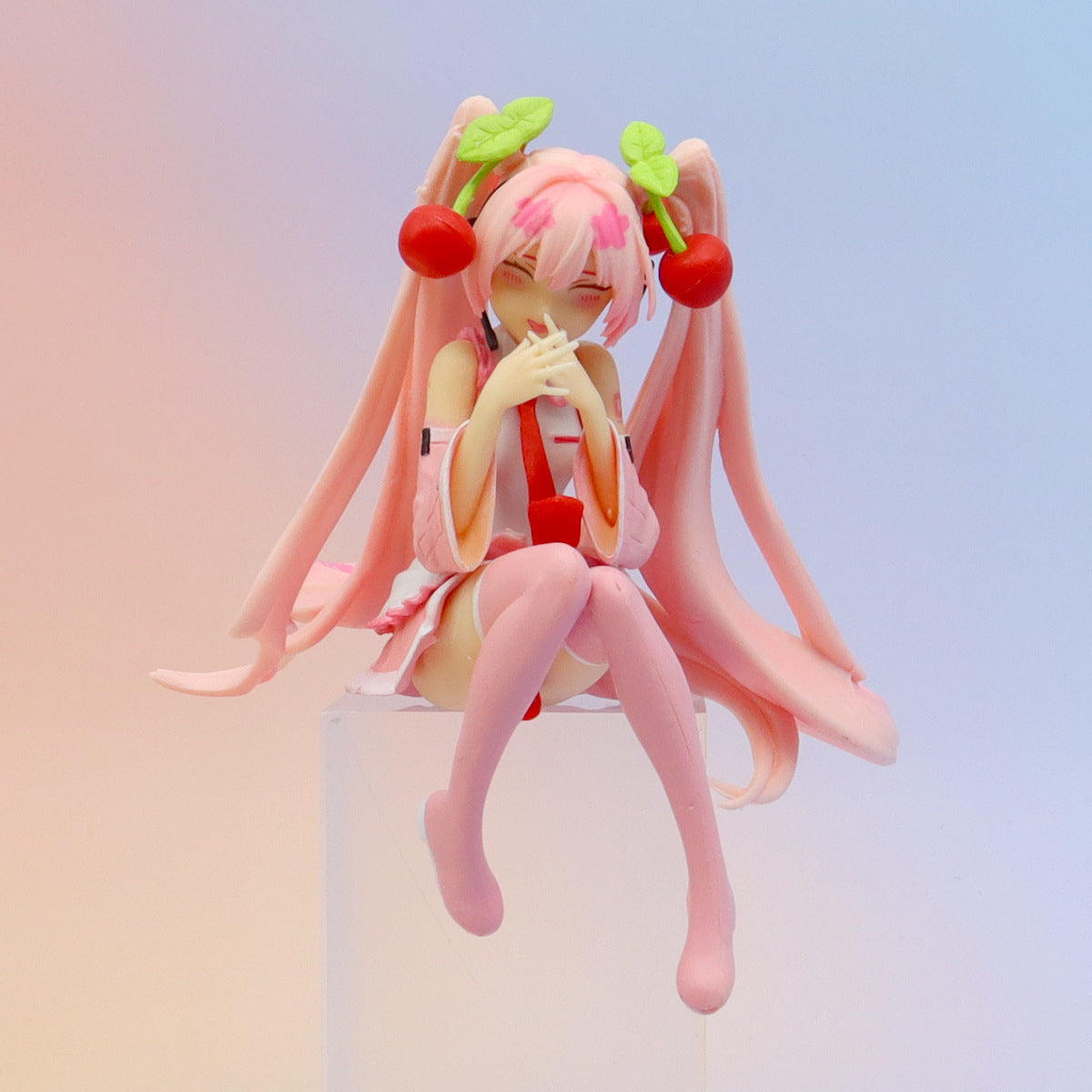 The Hatsune Miku Cherry Sitting Pose 11cm PVC Figure features a smiling character with long pink hair, cherry accessories, and knee-high socks. She wears a pink and white outfit and is seated gracefully against a soft gradient backdrop, hands gently near her face.