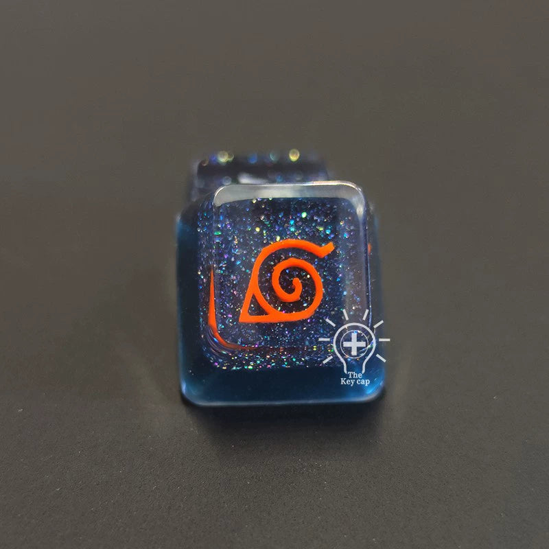 A close-up of a glittery dark blue resin keycap from the Naruto Symbols Keycap Set – Hidden Village, Uchiha, Akatsuki reveals an orange spiral leaf symbol. The sleek black background enhances its illuminated effect.