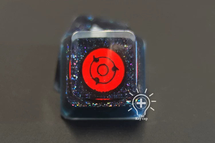 The &quot;Naruto Symbols Keycap Set – Hidden Village, Uchiha, Akatsuki&quot; by Naruto features a red Naruto symbol on black with a clear glossy cover and iridescent glitter. A small logo adorns the bottom right corner.