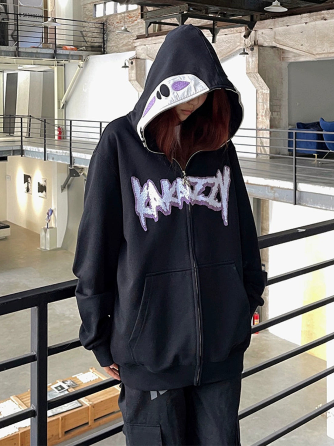 A person indoors wears a black No-Face Anime Hoodie by Spirited Away. The oversized zip-up has &quot;KARAZY&quot; in purple and white with an anime-inspired design. With the hood up, it partially obscures the face, set in a modern industrial space perfect for Spirited Away fans.