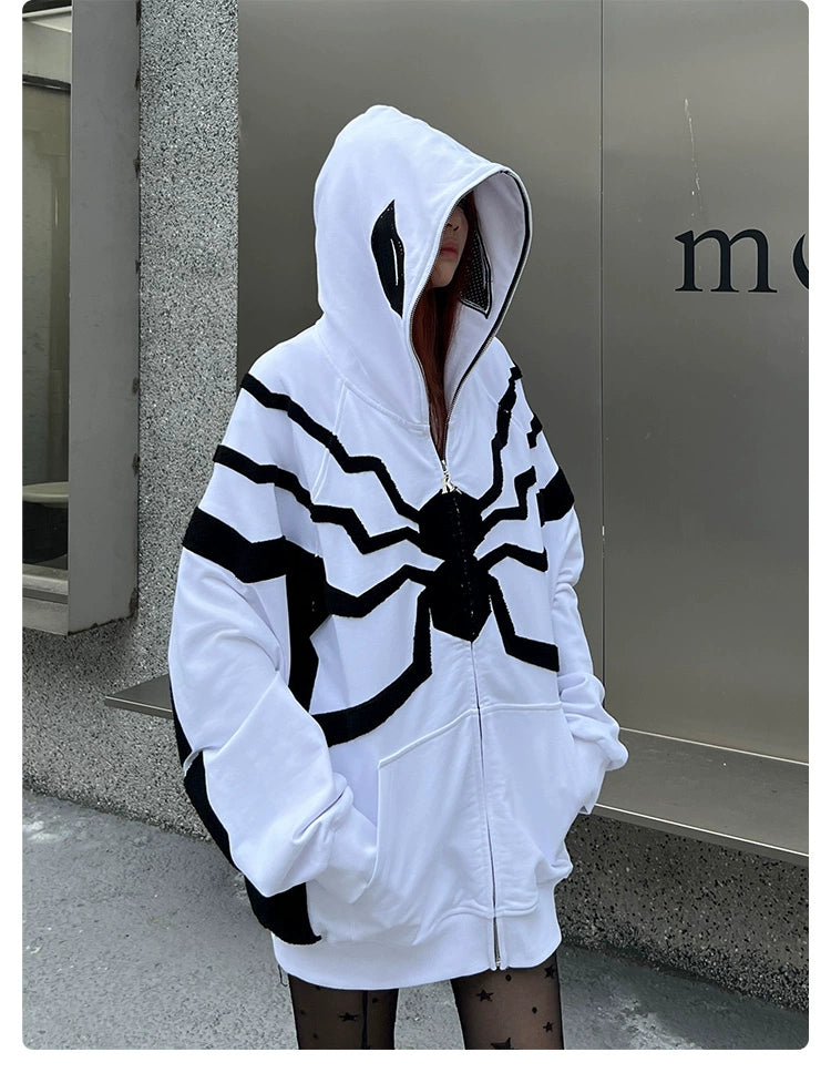 Against a concrete wall, someone stands with hands in pockets, wearing star-patterned leggings and a Seakoff Venom-Inspired White Hoodie featuring a black spider emblem. The 400GSM oversized zip-up superhero sweatshirt has its hood up, partially covering their face.