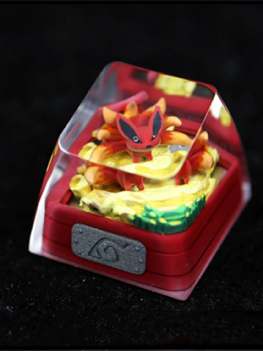 A small red fox figurine with multiple tails from the Bijuu collection is displayed in a clear case on a yellow and green swirl base. The case sits on a red square platform featuring a gray emblem inspired by Tailed Beasts Naruto Keycaps – Kurama, Shukaku, and Saiken by Naruto.