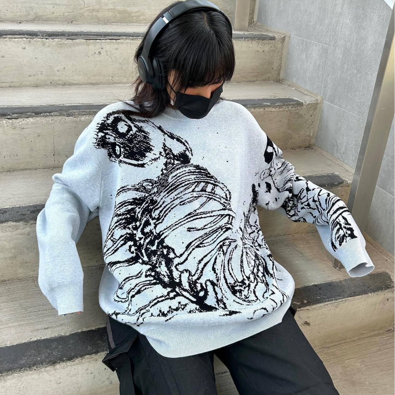 A person sits on concrete steps, sporting an alternative fashion style with Seakoff's Premium Skeleton Ribcage Knit Sweater in a gothic streetwear jacquard design. They pair it with black pants, a black face mask, large black headphones, and dark hair cascading as they rest their arms on their knees.