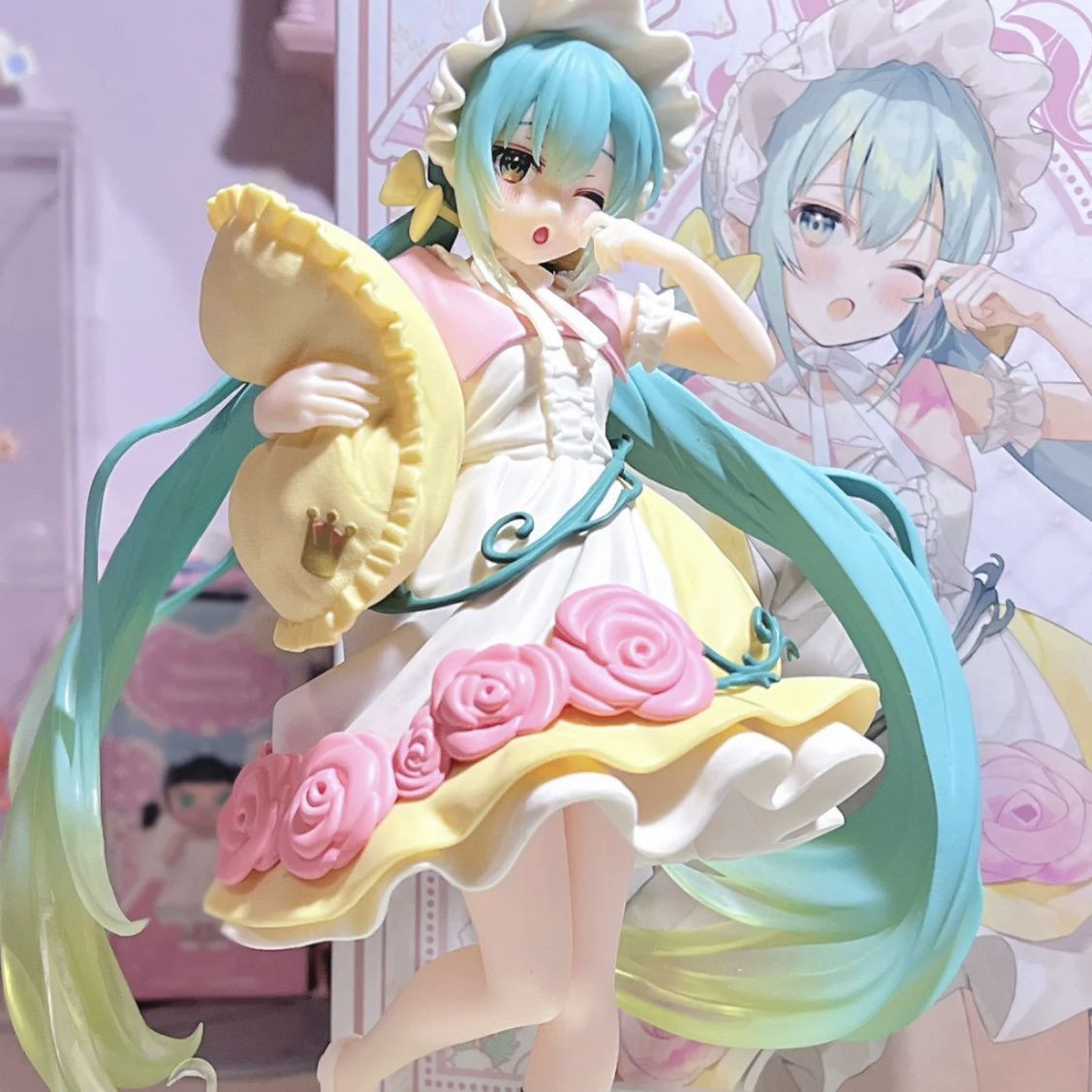 The Hatsune Miku Sleeping Beauty PVC Figure 20cm presents the iconic character with long blue-green hair, in a decorative outfit and bonnet, holding a pillow. Pink and yellow accents with rose patterns enhance the charming ensemble against its illustrated backdrop.