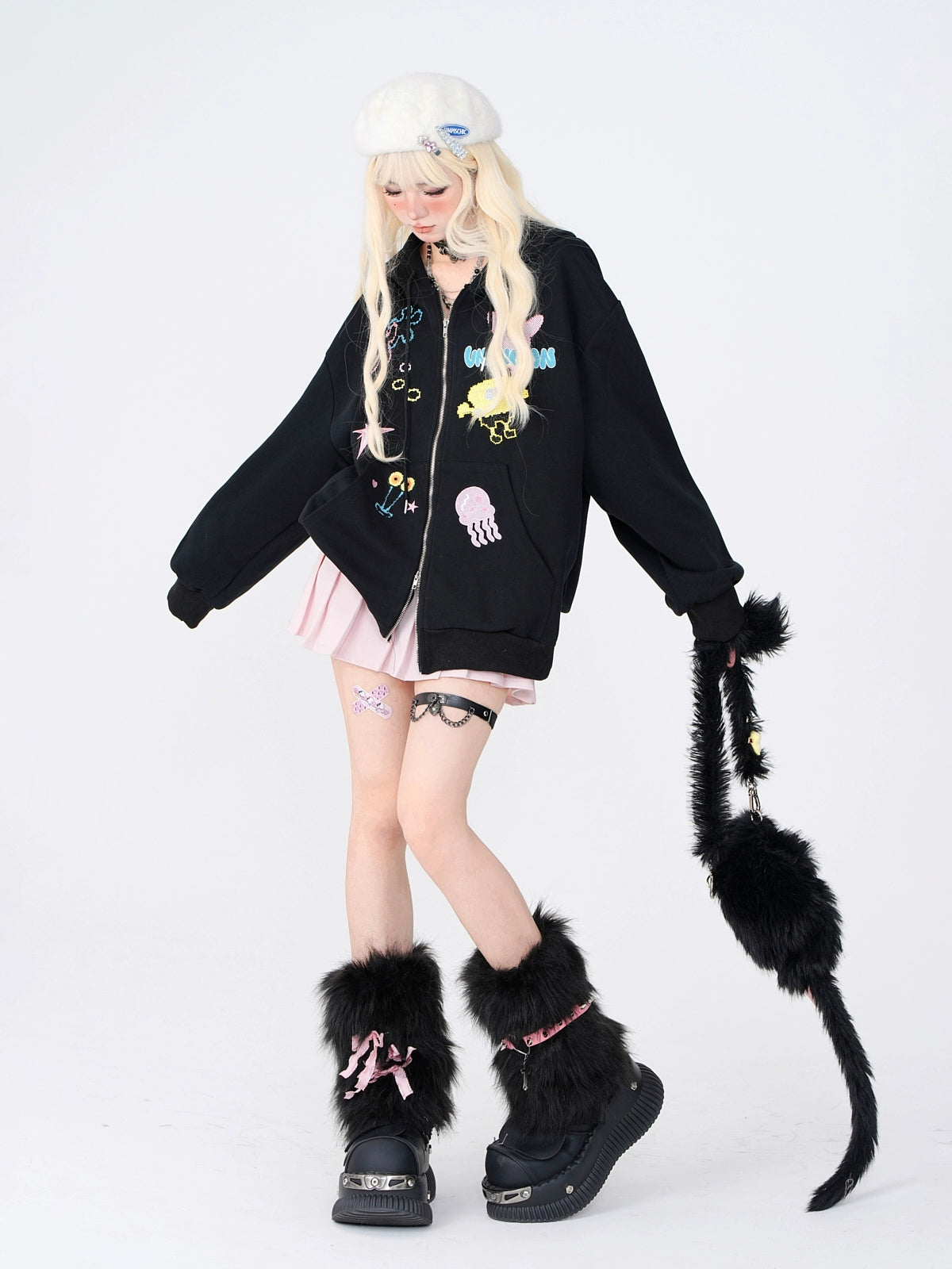 A person in a white beret, black jacket with colorful designs, pink skirt, and black furry boots poses against a plain backdrop. Holding a black furry bag with a tail-like extension, their style resembles Japanese streetwear vibes. Brand: Seakoff; Product: Kelly Kitty Pastel Kawaii Hoodie.