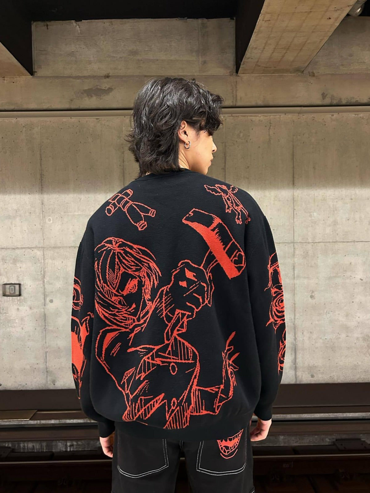 A person with dark, wavy hair stands in an industrial room with a concrete wall, wearing a black Premium Jujutsu Kaisen-Inspired Knit Sweater featuring red Yuji Itadori &amp; Nobara Kugisaki Jacquard Design by Jujutsu Kaisen.