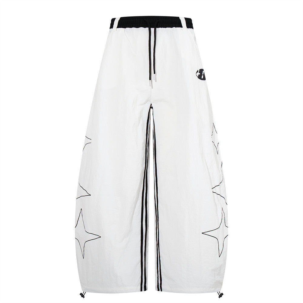 White wide-leg parachute pants with four-point star outline print, black elasticated waistband, center stripe detail, and dra