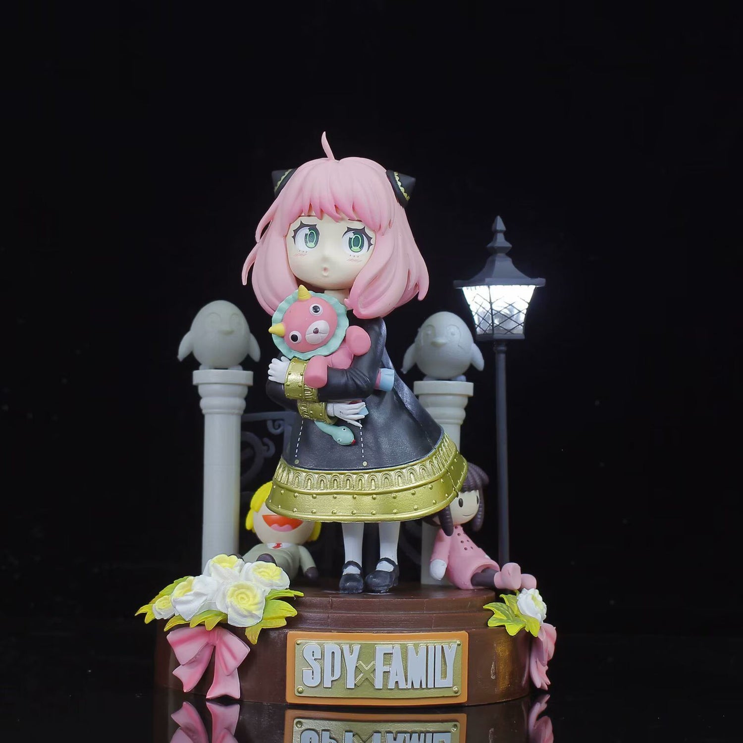 The Spy x Family Anya Forger PVC Figure features a 19 cm tall collectible of the pink-haired character in a black, gold-trimmed dress. She holds a plush toy and stands on a themed base with stone pillars, streetlamp, and flowers—ideal for anime collectibles fans.