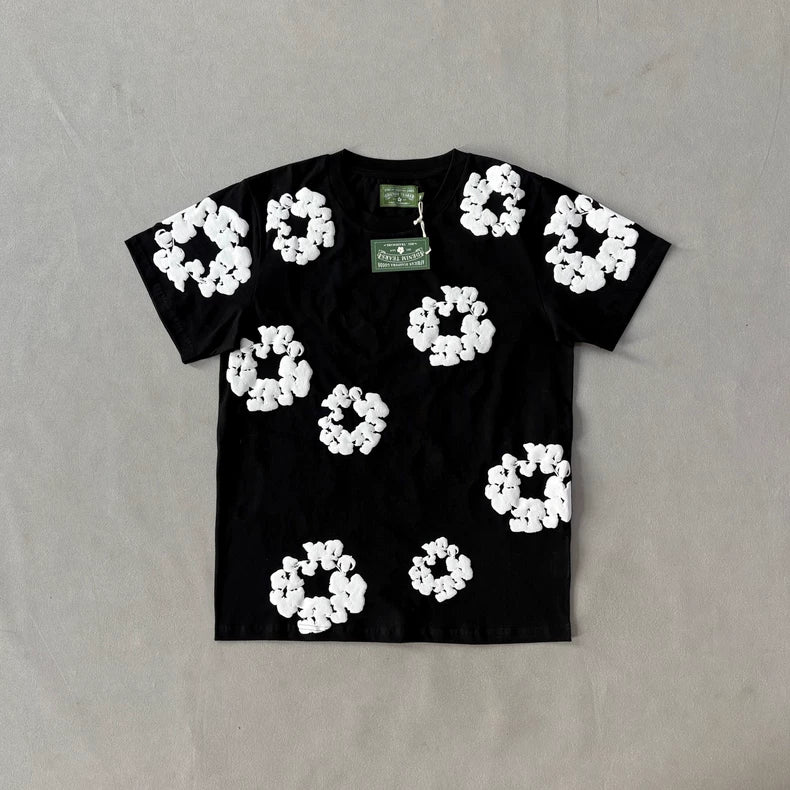 The Denim Tears Black Cotton Wreath All-Over Print T-Shirt features bold, oversized white floral graphic prints on the front, capturing striking streetwear style. Displayed against a light grey background.