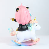 A Spy × Family Anya Forger PVC figure, 15 cm tall with pink hair and black attire, sits on a pastel-detailed rocking unicorn with a golden horn. The figure features an interchangeable face expression and points forward against a plain white background.