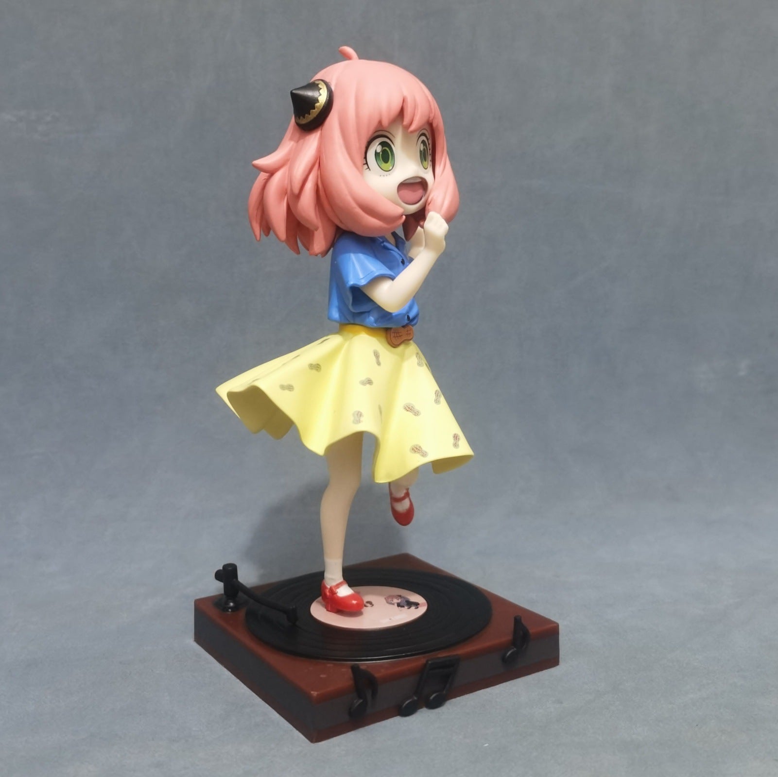 This limited edition Anya Forger Dancing Figure features the pink-haired Spy × Family character poised joyfully on one leg atop a record player, wearing a blue top and yellow skirt with dogs, and playfully posing with one hand near her mouth.