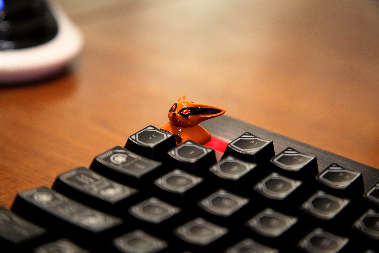 The Kurama Naruto Keycap, a chibi Nine-Tails Fox with black accents, is nestled among the keys of a black mechanical keyboard featuring Cherry MX switches. The keyboard is placed on a wooden surface against a blurred background.