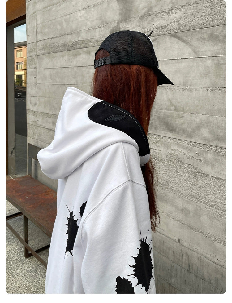 A person with long reddish-brown hair wears a Seakoff Monochrome Ink Splatter Hoodie, an oversized 400GSM white and black zip-up sweatshirt. They have a black cap on and are turned away on a wooden bench against a textured gray wall, embodying streetwear style.