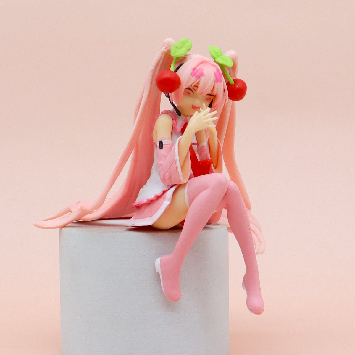 The Hatsune Miku Cherry Sitting Pose 11cm PVC figure features a smiling expression and cute design, with long pink hair and cherry-like accessories. It wears a pink and white outfit with matching boots, holds a drink cup atop a cylindrical pedestal, all set against a soft pink background.