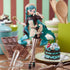 The Hatsune Miku Black Chocolate Pastry Chef 21cm PVC Figure, featuring teal hair, stands on the kitchen counter with chocolate bars, a whisk, a mixing bowl, and a cake. Cooking utensils complete this charming collectible scene.