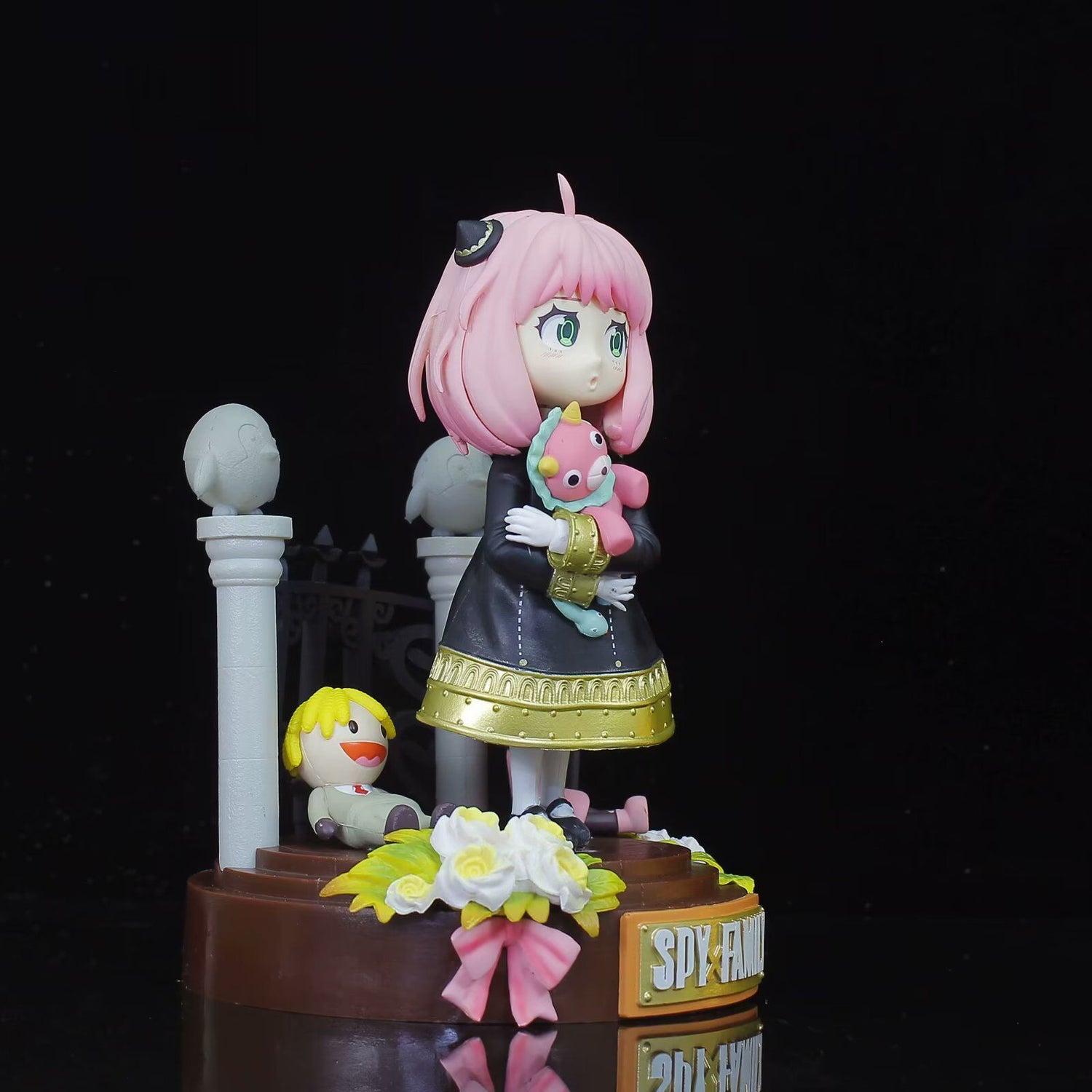 The Spy × Family Anya Forger PVC Figure is a 19 cm collectible statue featuring Anya in a black and gold dress with pink hair, holding a plush toy. She stands on a floral-adorned base with &quot;SPY FAMILY&quot; text, accompanied by a smiley face ball, decorative pillar, and gate—a must-have for anime fans.