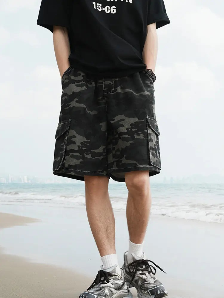 A person stands on the beach with hands in pockets, wearing a black T-shirt, Men's Baggy Camo Cargo Shorts with Elastic Drawstring Waist and Flap Pockets in black and gray camo, white socks, and gray sneakers. Ocean and cloudy sky in background.