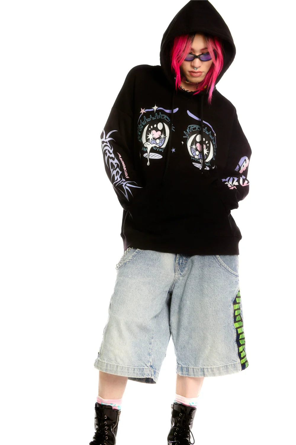 A person with pink hair wears a Seakoff Kawaii Aesthetic Crying Eyes Hoodie, styled in an anime design with colorful sleeve graphics. They're paired with sunglasses, oversized denim shorts, and black boots featuring pink laces, embodying streetwear fashion.
